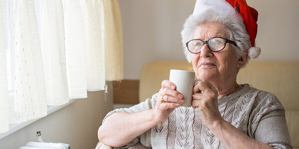 Senior woman with red Santa’s hat holding hot tea and looking through window. Xmas, home, age, alone, senior concept. Senior woman wearing santa hat