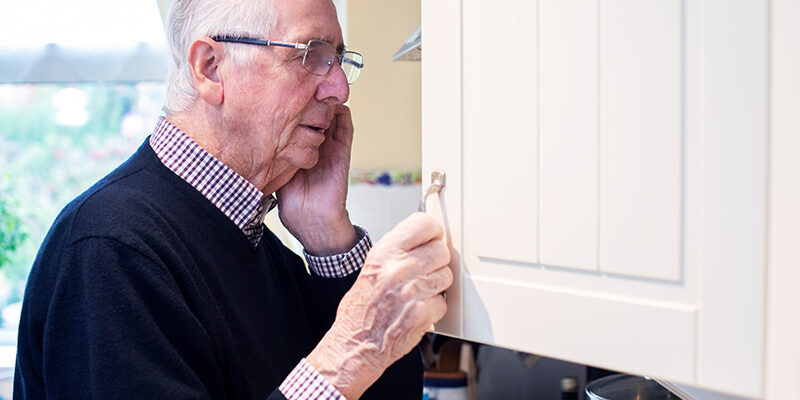 Forgetful Senior Man With Dementia Looking In Cupboard At Home