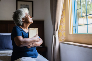 An older woman experiencing grief stares out the window while hugging a framed photo.